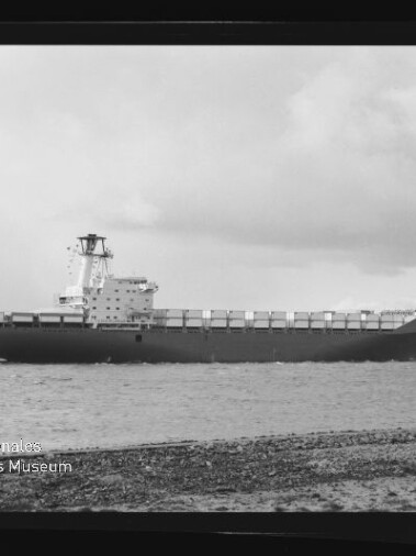 Schwarzweiß-Foto eines großen Containerschiffs, das nahe der Küste unter bewölktem Himmel fährt; im Vordergrund ein kiesiger Uferstreifen, auf dem Schiff mehrere Reihen Container und ein mittschiffs liegender Brückenaufbau, unten links das Logo des Internationalen Maritimen Museums Hamburg.