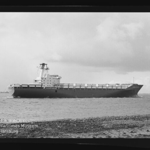 Schwarzweiß-Foto eines großen Containerschiffs, das nahe der Küste unter bewölktem Himmel fährt; im Vordergrund ein kiesiger Uferstreifen, auf dem Schiff mehrere Reihen Container und ein mittschiffs liegender Brückenaufbau, unten links das Logo des Internationalen Maritimen Museums Hamburg.