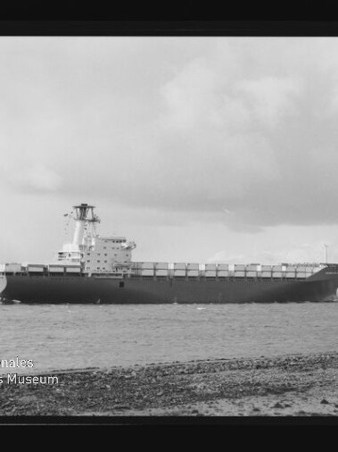 Schwarzweißaufnahme eines großen Containerschiffs in Seitenansicht auf ruhiger See, im Vordergrund ein Kiesstrand mit zwei kleinen Personen rechts, wolkiger Himmel und links unten das Signet Internationales Maritimes Museum Hamburg.