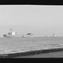 Schwarzweißaufnahme eines großen Frachtschiffs auf ruhiger See, begleitet von zwei Schleppern rechts, mit Kai im Vordergrund und Wasserzeichen des Internationalen Maritimen Museums Hamburg unten links; Filmbandrand sichtbar.