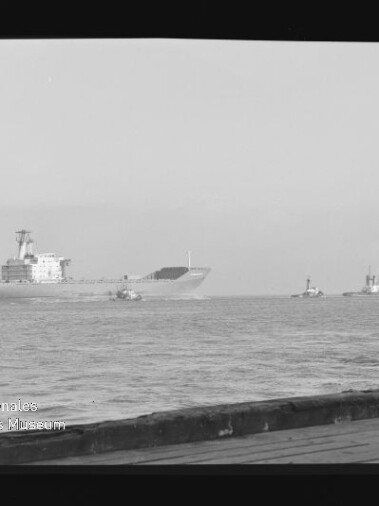 Schwarzweißaufnahme eines großen Frachtschiffs auf ruhiger See, begleitet von zwei Schleppern rechts, mit Kai im Vordergrund und Wasserzeichen des Internationalen Maritimen Museums Hamburg unten links; Filmbandrand sichtbar.