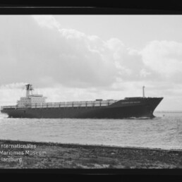 Schwarz-weiße Aufnahme eines langen Containerschiffs in Seitenansicht, das nahe der Küste auf ruhiger See unter einem wolkenverhangenen Himmel fährt.