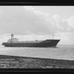 Schwarzweißfoto eines großen Frachtschiffs mit auf Deck gestapelten Containern, das nahe der Küste über ruhiges Wasser fährt; im Vordergrund ein schmaler Uferstreifen, im Hintergrund wolkenverhangener Himmel und unten links ein Museumslogo.
