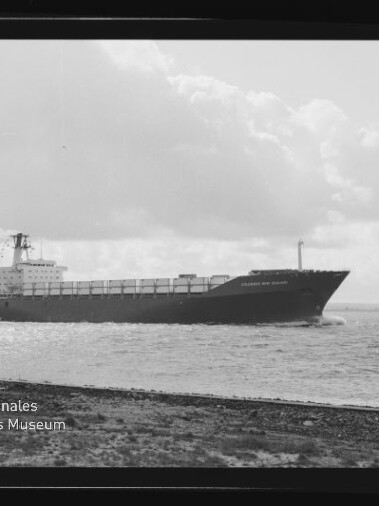 Schwarzweißfoto eines großen Frachtschiffs mit auf Deck gestapelten Containern, das nahe der Küste über ruhiges Wasser fährt; im Vordergrund ein schmaler Uferstreifen, im Hintergrund wolkenverhangener Himmel und unten links ein Museumslogo.
