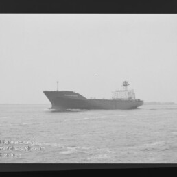 Schwarzweißaufnahme eines großen Frachtschiffs, das auf ruhiger See nahe dem Horizont fährt, mit sichtbarem Filmrand (AGFA PAN 400) und Museumswasserzeichen unten links.