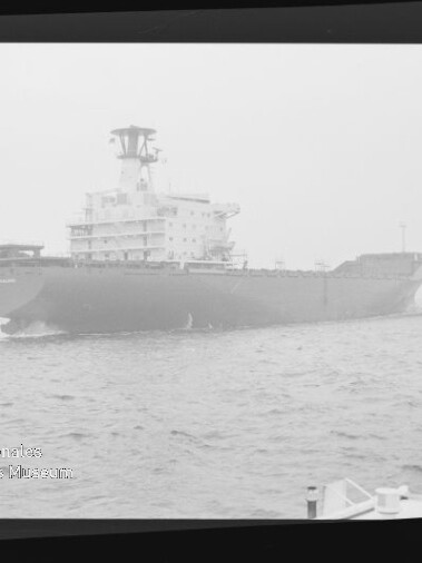 Großes Frachtschiff in diesiger See, aufgenommen schwarzweiß innerhalb eines Filmnegativrahmens mit sichtbaren Randmarkierungen; im Vordergrund rechts Teile des Decks und ein Poller.