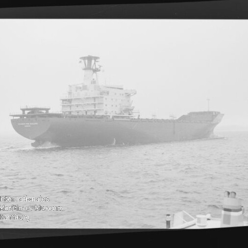Großes Frachtschiff in diesiger See, aufgenommen schwarzweiß innerhalb eines Filmnegativrahmens mit sichtbaren Randmarkierungen; im Vordergrund rechts Teile des Decks und ein Poller.