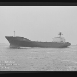 Schwarzweißseitaufnahme eines großen Frachtschiffs mit hohem Aufbaudeck am Heck, das auf ruhiger See fährt; sichtbare Filmborder und Wasserzeichen des Internationalen Maritimen Museums Hamburg.