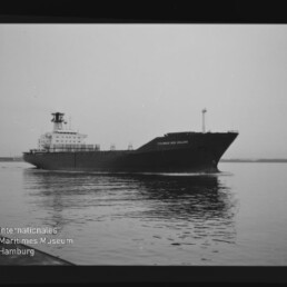 Schwarzweißaufnahme eines Frachtschiffs mit markantem Bug und achterlichem Aufbau, das ruhig über spiegelndes Wasser fährt; links unten Stempel des Internationalen Maritimen Museums Hamburg.