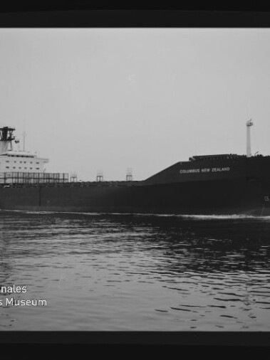Schwarzweißaufnahme des Frachtschiffs Columbus New Zealand, seitlich auf ruhigem Wasser fahrend, mit Aufbau und Schornstein links sowie dem Schriftzug und Logo des Internationalen Maritimen Museums Hamburg unten links.