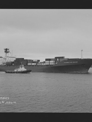 Schwarz-weiß-Foto eines beladenen Containerschiffs „Columbus New Zealand“ auf ruhigem Wasser, begleitet von einem Schlepper, mit gestapelten Containern, niedrigem Horizont, sichtbarem Filmrand (Kodak Safety Film) und Museumswasserzeichen.