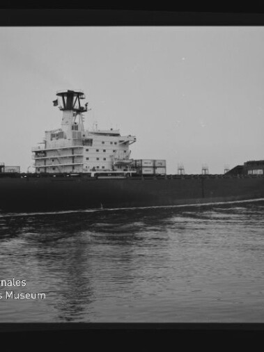 Schwarzweißaufnahme eines großen Frachtschiffs in Seitenansicht auf ruhigem Wasser, mit markantem Brückenaufbau und vereinzelten Containern auf dem Deck; links unten ist ein Museumswasserzeichen sichtbar.