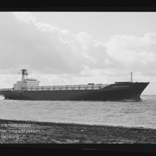 Schwarzweiß-Foto eines großen Frachtschiffs in Seitenansicht, das bei ruhiger See nahe der Küste unter einem wolkigen Himmel fährt, unten links sichtbar das Signet des Internationalen Maritimen Museums Hamburg.