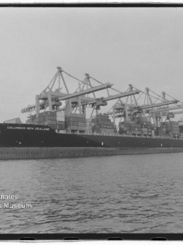 Schwarzweißaufnahme eines großen Containerschiffs 'COLUMBUS NEW ZEALAND' am Kai, beladen mit gestapelten Containern und umgeben von mehreren großen Hafenkränen, Wasser im Vordergrund und Museumslogo unten links.