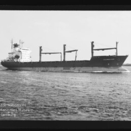 Schwarzweiß-Foto eines großen Frachtschiffs mit drei Ladekranen, das bei ruhiger See parallel zum Bildrand fährt; unten links ein Wasserzeichen des Internationalen Maritimen Museums Hamburg.