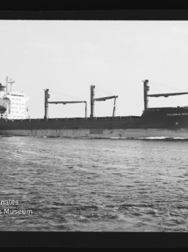 Schwarzweiß-Foto eines großen Frachtschiffs mit drei Ladekranen, das bei ruhiger See parallel zum Bildrand fährt; unten links ein Wasserzeichen des Internationalen Maritimen Museums Hamburg.