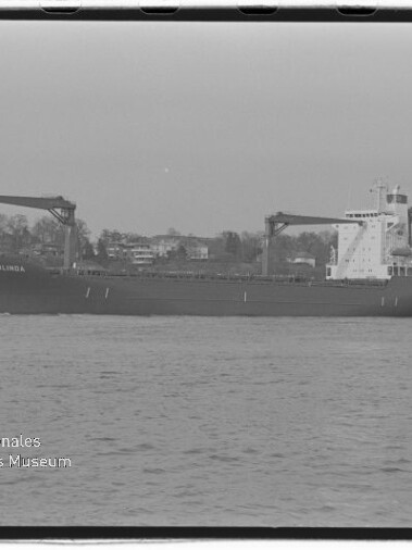 Schwarzweiß-Foto eines Frachtschiffs „COLUMBUS OLINDA“ mit zwei Ladekränen auf dem Wasser vor einem Ufer mit Häusern und Bäumen, unten links das Logo des Internationalen Maritimen Museums Hamburg sichtbar.
