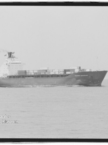 Schwarzweiß-Foto eines großen Containerschiffs in Seitenansicht auf ruhiger See mit gestapelten Containern an Deck, leichtem Rauch aus dem Schornstein und Wasserzeichen des Internationalen Maritimen Museums Hamburg unten links.