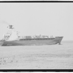 Schwarzweiß-Foto eines Containerschiffs auf ruhiger See mit gestapelten Containern, sichtbaren Aufbauten und leichter Bugwelle; links unten ist das Wasserzeichen des Internationalen Maritimen Museums Hamburg zu sehen.