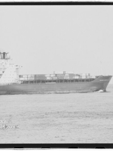 Schwarzweiß-Foto eines Containerschiffs auf ruhiger See mit gestapelten Containern, sichtbaren Aufbauten und leichter Bugwelle; links unten ist das Wasserzeichen des Internationalen Maritimen Museums Hamburg zu sehen.