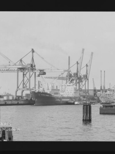 Schwarzweißaufnahme eines Industriehafens mit mehreren großen Kranen am Kai, einem festgemachten Frachtschiff in der Bildmitte, Hafenanlagen und Pollern im Vordergrund sowie dem Wasserzeichen ‚Internationales Maritimes Museum Hamburg‘ unten links.