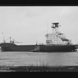 Schwarz-weiß-Aufnahme eines großen Frachtschiffs mit hohem Brückenturm, begleitet von einem Schlepper auf dem Wasser, Hafenbauten im Hintergrund und dem Logo des Internationalen Maritimen Museums Hamburg in der linken unteren Ecke.