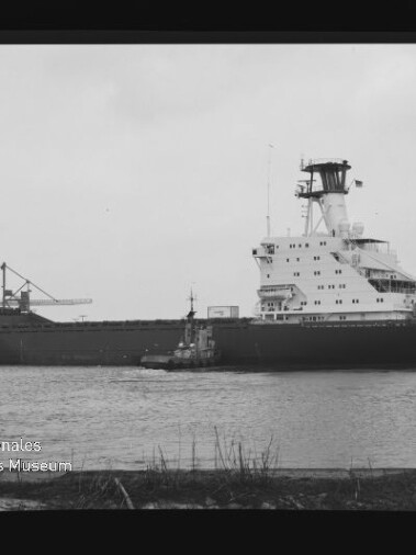 Schwarz-weiß-Aufnahme eines großen Frachtschiffs mit hohem Brückenturm, begleitet von einem Schlepper auf dem Wasser, Hafenbauten im Hintergrund und dem Logo des Internationalen Maritimen Museums Hamburg in der linken unteren Ecke.