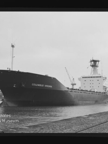 Schwarzweißaufnahme des Frachtschiffs Columbus Virginia, das an einem Industriehafen mit Kränen im Hintergrund am Kai liegt; im Vordergrund Schienen und ein Poller, unten links ein Museumswasserzeichen.