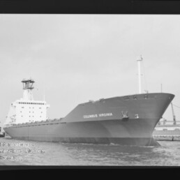 Schwarzweiß-Foto eines großen Frachtschiffs mit dem Namen COLUMBUS VIRGINIA, Bug vorn, im Hafen mit sichtbaren Kränen und Kai im Hintergrund und Wasser im Vordergrund.