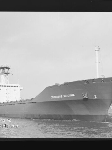 Schwarzweiß-Foto eines großen Frachtschiffs mit dem Namen COLUMBUS VIRGINIA, Bug vorn, im Hafen mit sichtbaren Kränen und Kai im Hintergrund und Wasser im Vordergrund.
