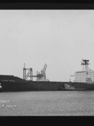 Schwarzweiß-Aufnahme eines Frachtschiffs Columbus Virginia im Hafen, seitliche Ansicht mit zwei Schleppern am Bug, Hafenkränen im Hintergrund und Wasserzeichen des Internationalen Maritimen Museums Hamburg links unten.