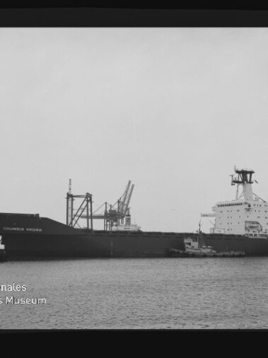 Schwarzweiß-Aufnahme eines Frachtschiffs Columbus Virginia im Hafen, seitliche Ansicht mit zwei Schleppern am Bug, Hafenkränen im Hintergrund und Wasserzeichen des Internationalen Maritimen Museums Hamburg links unten.