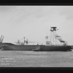Schwarzweißaufnahme eines großen Frachtschiffs mit achterlichem Aufbauten und einem Schlepper neben dem Rumpf, vor Hafenkränen auf ruhigem Wasser, eingefasst von einem Filmrand mit AGFAPAN-Kennung und Museumslogo.