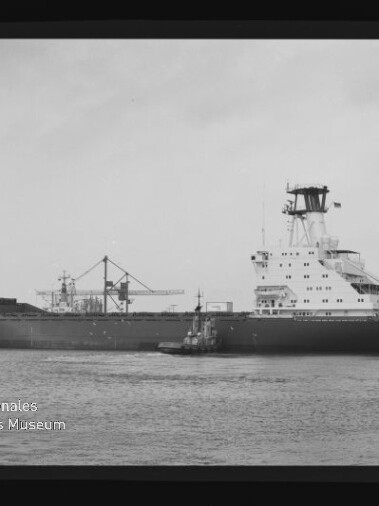 Schwarzweißaufnahme eines großen Frachtschiffs mit achterlichem Aufbauten und einem Schlepper neben dem Rumpf, vor Hafenkränen auf ruhigem Wasser, eingefasst von einem Filmrand mit AGFAPAN-Kennung und Museumslogo.