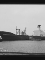Schwarzweißaufnahme eines großen Frachtschiffs namens „Columbus Virginia“ im Hafen, von einem Schlepper begleitet, vor Kaianlagen und Ladekränen; Museumslogo unten links.