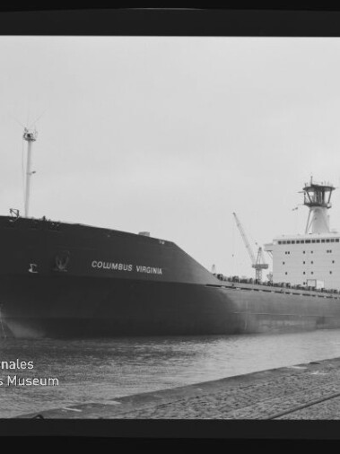 Großes Frachtschiff „COLUMBUS VIRGINIA“ in Schwarz-Weiß, am Kai eines Industriehafens festgemacht mit sichtbarem Schiffsrumpf, Kranen im Hintergrund und Wasserzeichen des Internationalen Maritimen Museums Hamburg.