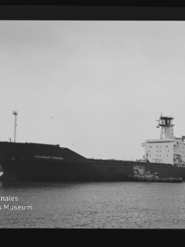 Schwarzweißfoto eines großen Frachtschiffs COLUMBUS VIRGINIA im Hafen, ein Schlepper am Heck, Hafenkräne im Hintergrund, Negativrahmen mit Nummer links und Museumssignet unten links.