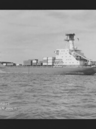 Schwarzweiß-Foto einer Seitenansicht eines Containerschiffs mit gestapelten Containern und markantem Brückenaufbau, das auf ruhiger See vor einer flachen Küstenlinie fährt; sichtbarer Filmrand und Wasserzeichen 'Internationales Maritimes Museum Hamburg' unten links.