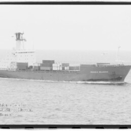 Schwarzweiß-Foto eines Containerschiffs Columbus Wellington mit gestapelten Containern, das bei ruhiger See nach rechts fährt; links unten ein rundes Museumswasserzeichen.