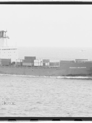 Schwarzweiß-Foto eines Containerschiffs Columbus Wellington mit gestapelten Containern, das bei ruhiger See nach rechts fährt; links unten ein rundes Museumswasserzeichen.