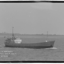 Schwarzweiß-Foto eines Frachtschiffs mit dem Namen Colonia, das ruhig in Küstennähe fährt; im Vordergrund leicht welliges Meer und unten links das Logo sowie der Schriftzug des Internationalen Maritimen Museums Hamburg.
