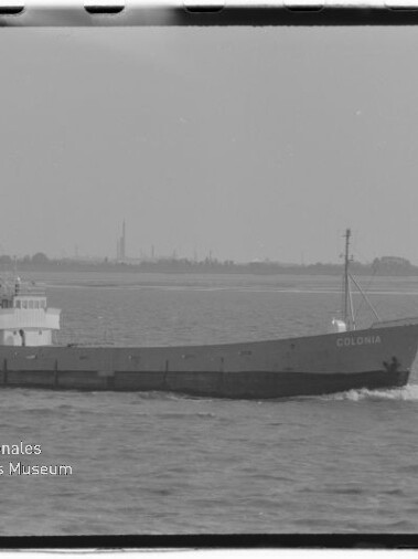 Schwarzweiß-Seitenansicht eines kleinen Frachtschiffs mit Namen COLONIA, achtern liegendem Aufbau und einzelnem Mast, das mit leichter Bugwelle auf ruhiger See vor einer flachen, industriell geprägten Küstenlinie fährt.