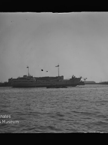 Schwarzweiß-Foto eines großen Passagier- oder Fährschiffs mit gehissten Flaggen auf ruhigem Hafenwasser, im Hintergrund Silhouetten von Landanlagen und markanten Türmen; links unten sichtbares Wasserzeichen des Internationalen Maritimen Museums Hamburg sowie altersbedingte Flecken und Kratzer im Negativ.