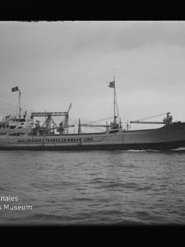 Schwarzweiß-Foto eines Schiffs mit auf dem Rumpf sichtbarer Aufschrift 'Trans-Caribbean Line', Ladekränen und Flaggen, das an einem Industriehafen mit großem Portalkran vorbeifährt; Museumswasserzeichen unten links.