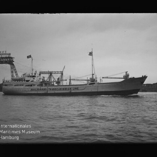 Schwarzweiß-Foto eines Schiffs mit auf dem Rumpf sichtbarer Aufschrift 'Trans-Caribbean Line', Ladekränen und Flaggen, das an einem Industriehafen mit großem Portalkran vorbeifährt; Museumswasserzeichen unten links.
