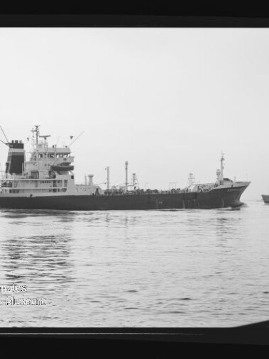 Schwarzweißaufnahme eines großen Tankers auf ruhiger See mit einer kleinen Begleitbarkasse rechts, gezeigt innerhalb eines Negativrahmens mit Filmkennzeichnung ‚AGFAPAN 100‘ und sichtbarem Museumswasserzeichen unten links.