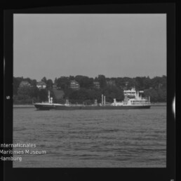 Schwarzweißaufnahme eines Frachtschiffs, das auf ruhigem Wasser vor einem baumbestandenen Ufer mit einzelnen Häusern fährt; umlaufender Filmrand und das Logo des Internationalen Maritimen Museums Hamburg sind links unten sichtbar.