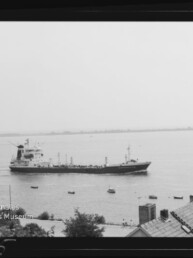 Schwarzweißaufnahme eines Frachtschiffs, das nahe der Küste auf ruhigem Gewässer fährt; mehrere kleine Boote umgeben das Schiff, im Vordergrund sind Hausdächer und Vegetation zu sehen und links unten ein Museumslogo.