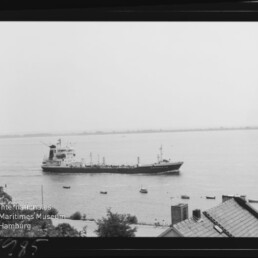 Schwarzweißaufnahme eines Frachtschiffs, das nahe der Küste auf ruhigem Gewässer fährt; mehrere kleine Boote umgeben das Schiff, im Vordergrund sind Hausdächer und Vegetation zu sehen und links unten ein Museumslogo.