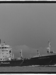 Schwarz‑weiß-Foto eines Frachtschiffs namens COMANCHE, seitliche Ansicht auf See; links unten Wasserzeichen des Internationalen Maritimen Museums Hamburg.